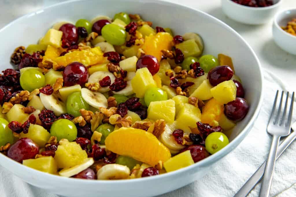 A bowl of fresh fruit salad featuring green and red grapes, orange slices, banana slices, pineapple chunks, and dried cranberries. The salad is topped with chopped nuts, and a fork is placed beside the bowl on a white surface.