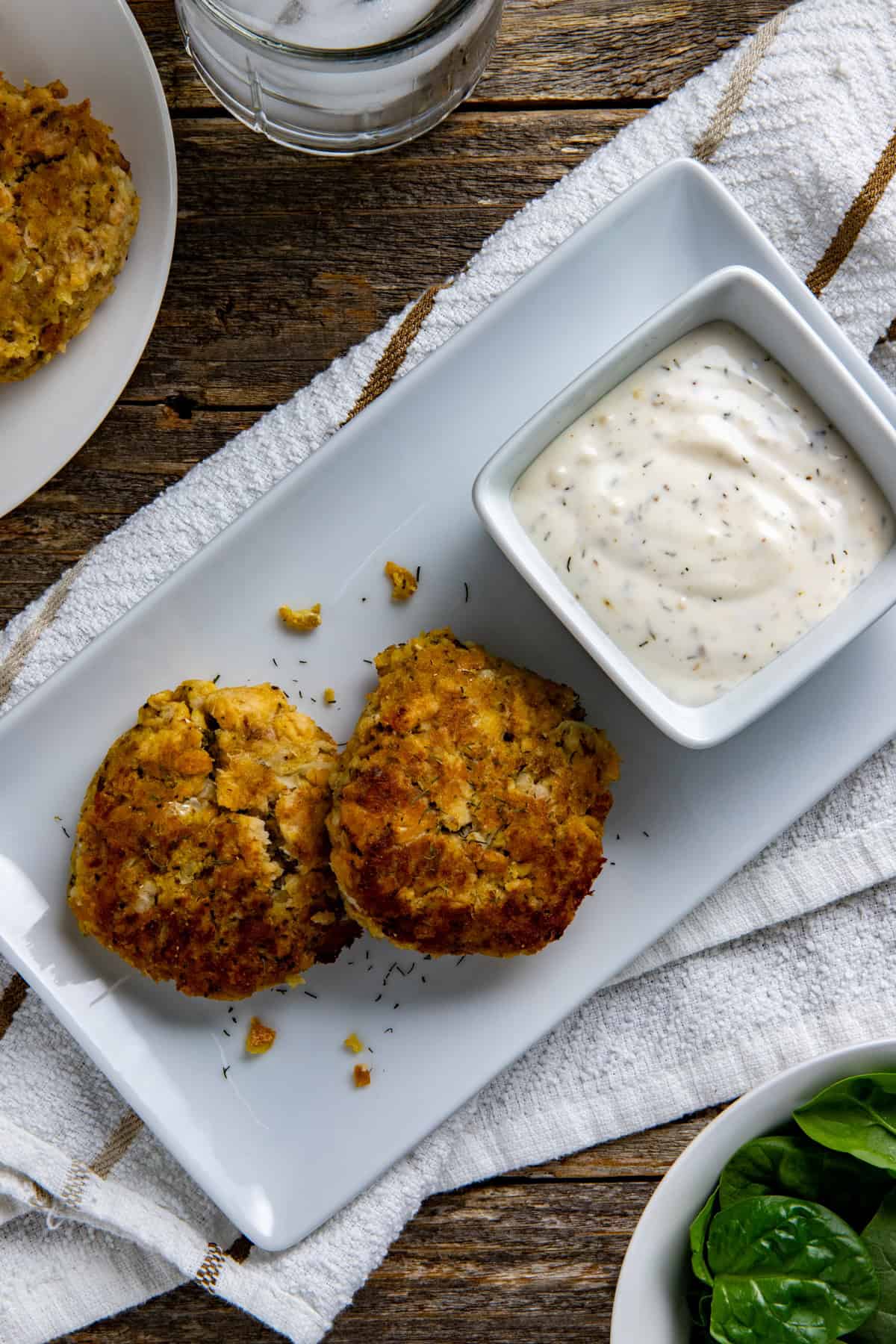 Two cooked salmon patties on a white serving tray with a square bowl of lemon dill sauce.