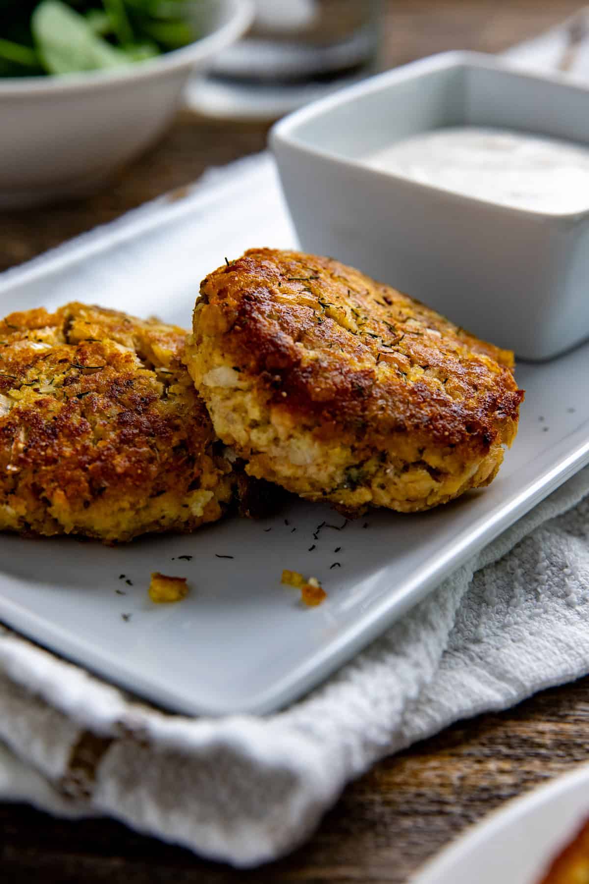 Two cooked salmon patties on a white serving tray with a square bowl of lemon dill sauce.