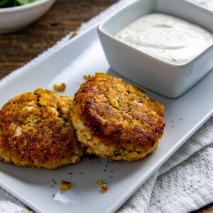 Two cooked salmon patties on a white serving tray with a square bowl of lemon dill sauce.