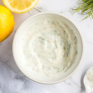 Blended lemon dill sauce in white bowl with sliced lemons on the side.