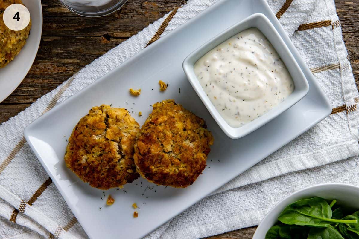 Two cooked salmon patties on a white serving tray with a square bowl of lemon dill sauce.