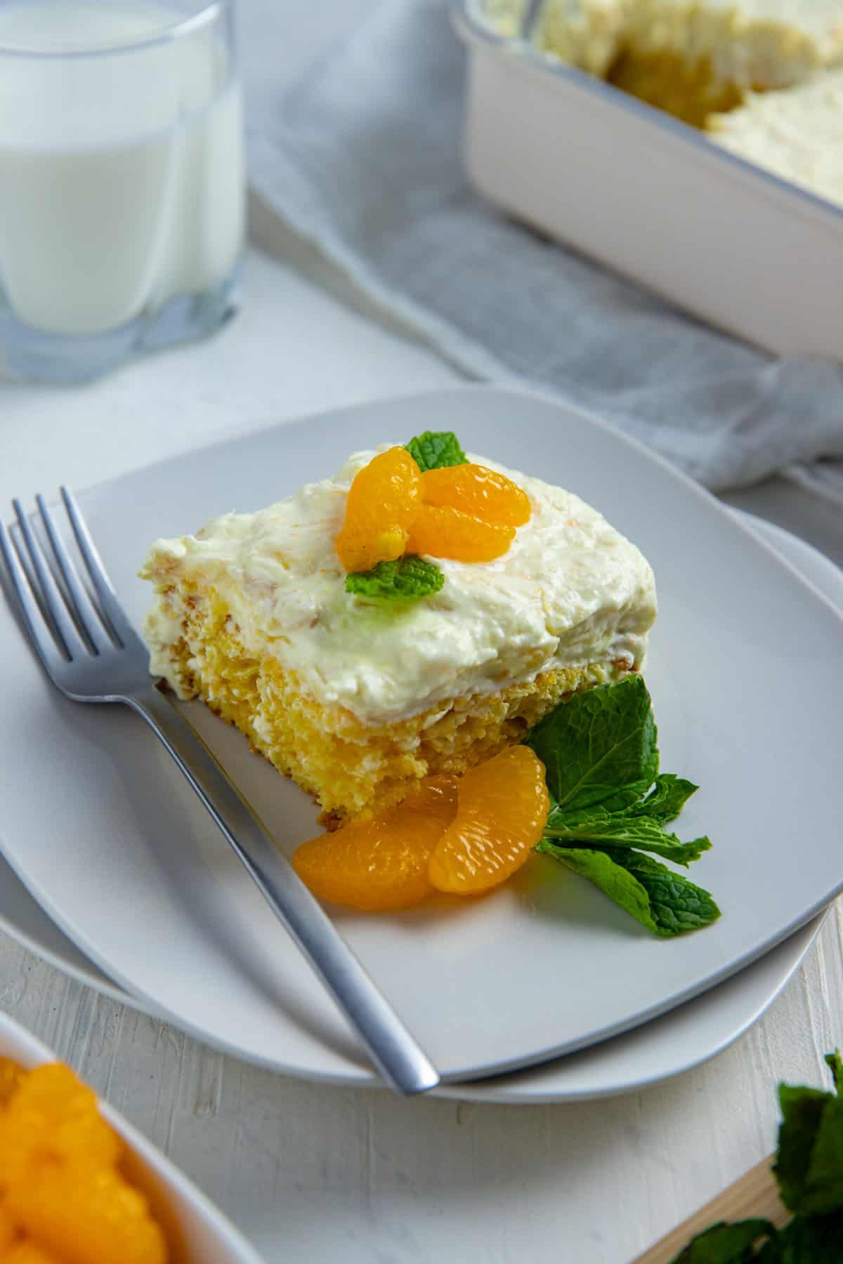 A slice of cake topped with a couple of mandarin orange segments and mint leaves on a square white plate with a fork.