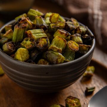 Roasted sliced okra piled high in a brown bowl on a wooden serving platter with a spoon on the side.