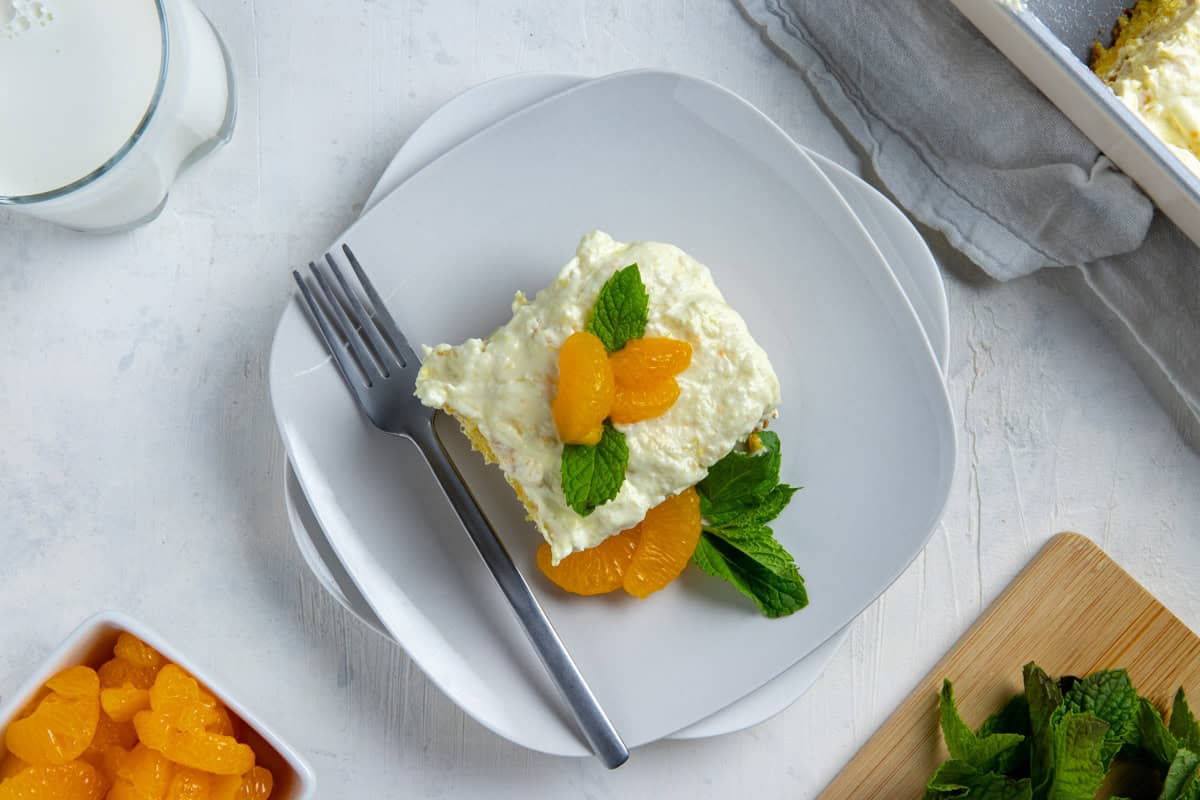A slice of cake topped with a couple of mandarin orange segments and mint leaves on a square white plate with a fork.