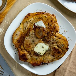 One cooked and seasoned pork chop with a pat of butter on top on a white serving plate.