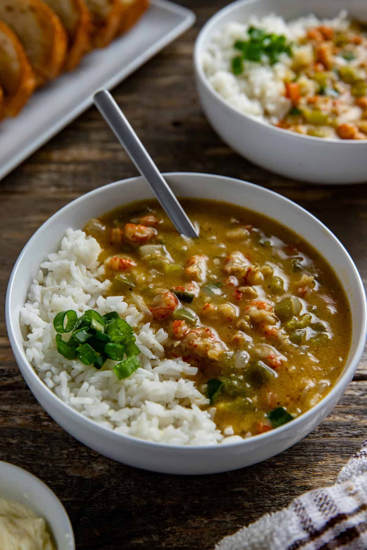 A bowl of etouffee with white rice. Sliced green onions for garnish. With sliced, toasted French bread in the background.