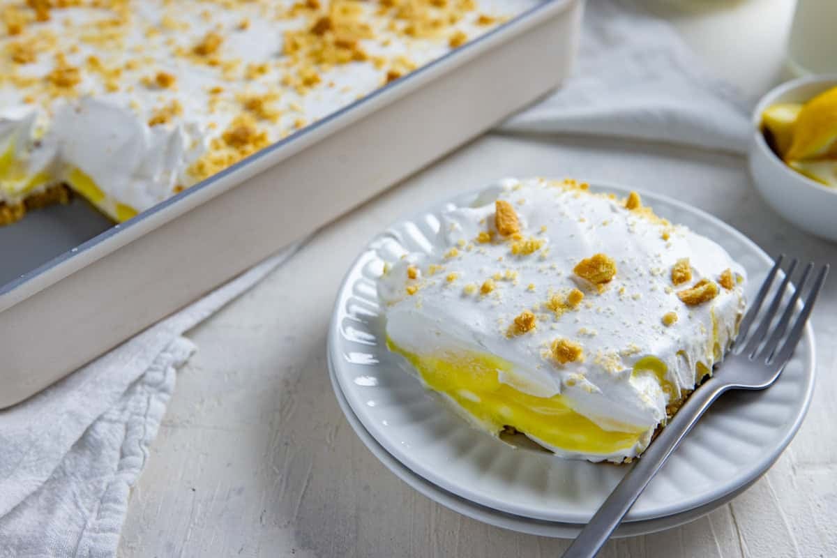 A serving of lemon lush on a white plate with a fork on the side. The pan of lemon lush is in the background along with a bowl of sliced lemon wedges.