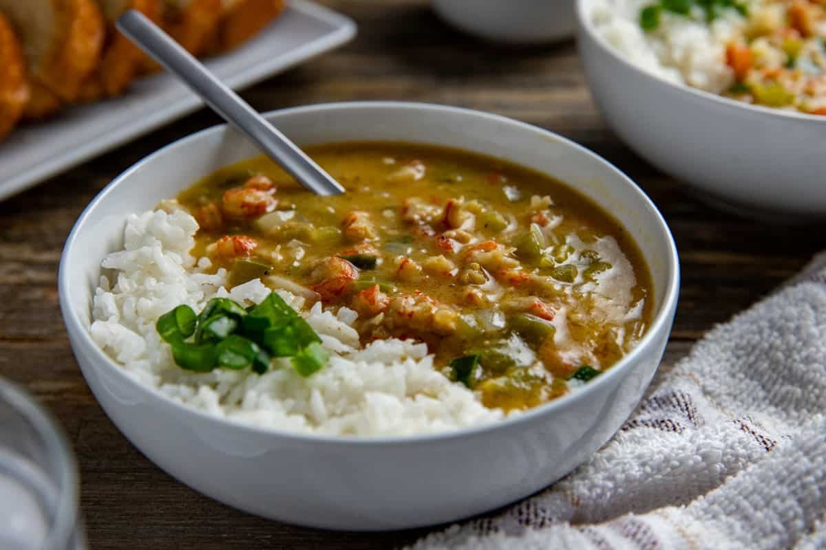 A bowl of etouffee with white rice. Sliced green onions for garnish. With sliced, toasted French bread in the background.