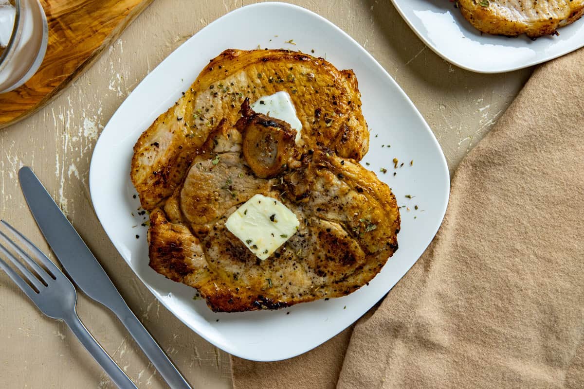 A cooked pork chop with a pat of butter and sprinkle of dried herbs on top in a white square plate with a fork and knife on the side.