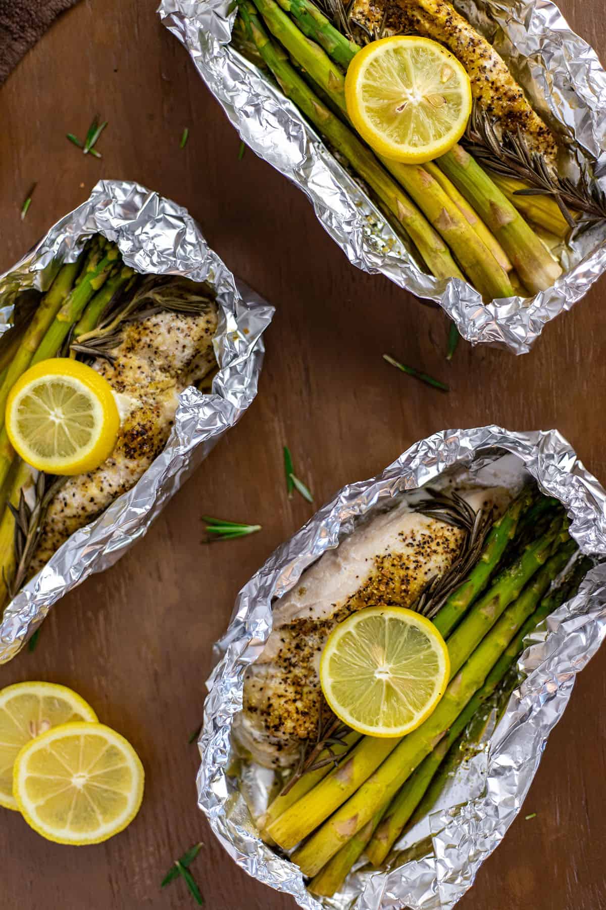 Three foil packets with baked chicken breast with asparagus spears, rosemary sprig, and lemon slice.