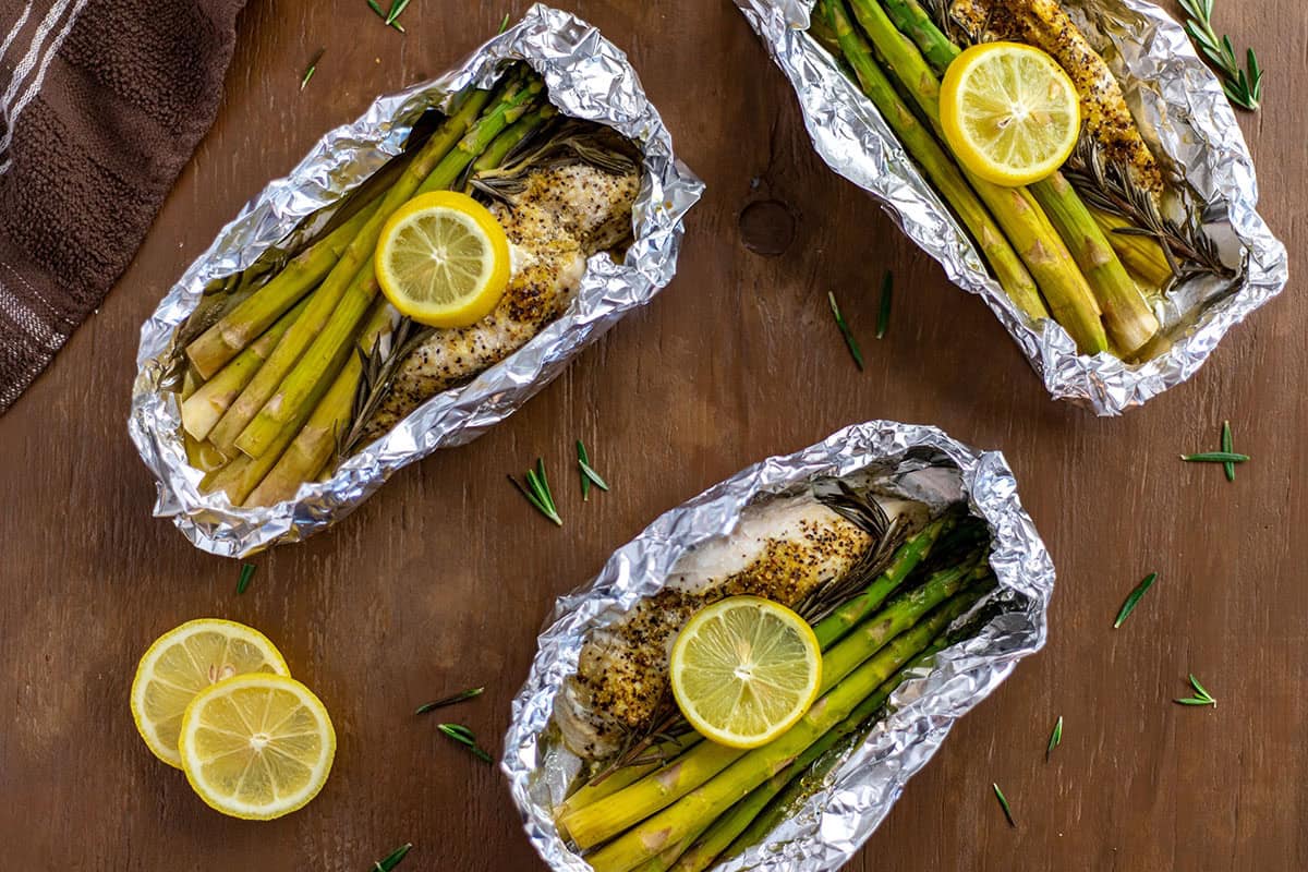 Three Lemon Pepper Chicken with rosemary and asparagus inside foil packet with lemon slices on top.