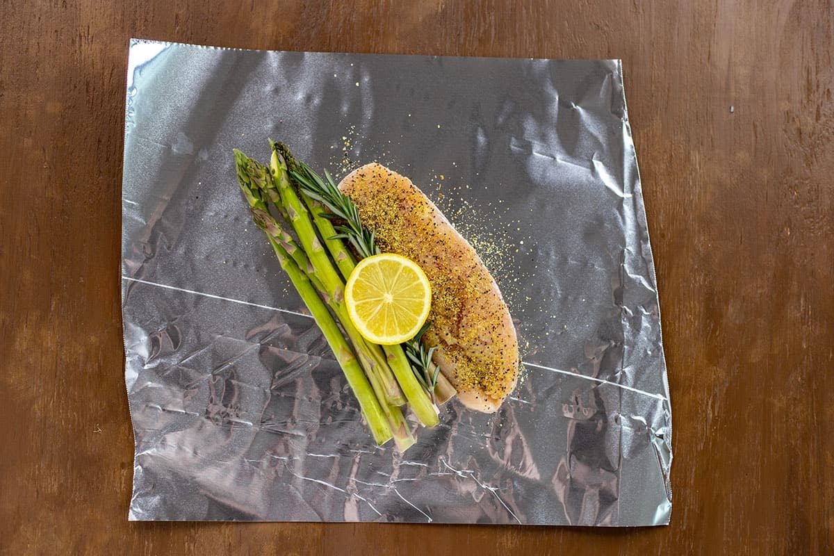 Uncooked seasoned chicken breast with asparagus, rosemary, and lemon slice on sheet of foil.