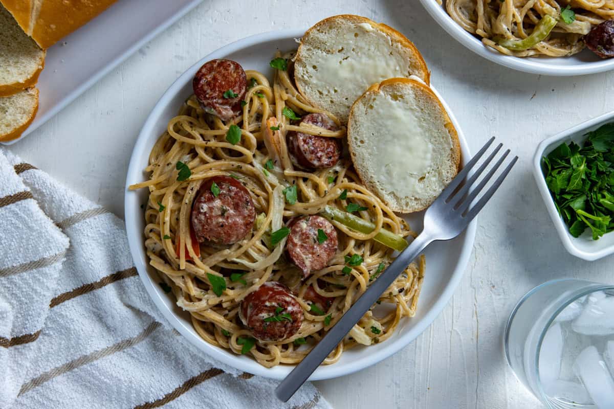 Cooked cajun sausage spaghetti on white plate with a fork and two slices of buttered French bread on the side.