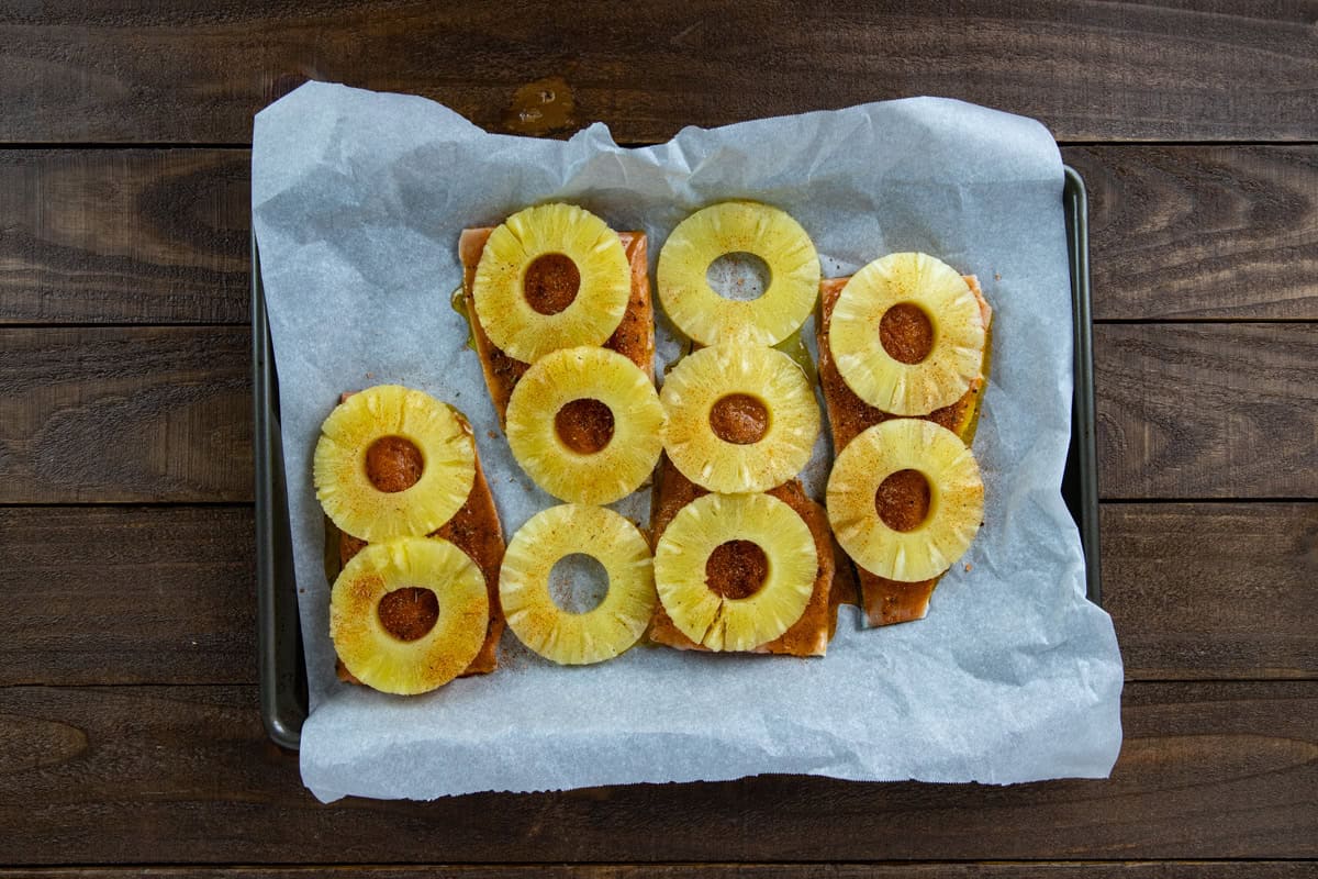Four uncooked salmon fillets on a parchment lined sheet pan. Topped with honey-olive oil mixture, seasonings, and pineapple rings.