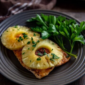 One baked salmon fillet topped with two pineapple rings and sprinkled with fresh cut parsley on a brown plate.