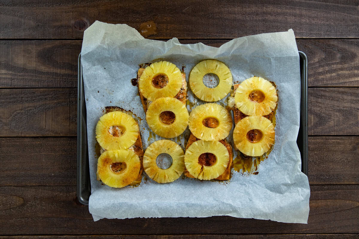 Four baked salmon fillets on a parchment lined sheet pan. Topped with honey-olive oil mixture, seasonings, and pineapple rings.