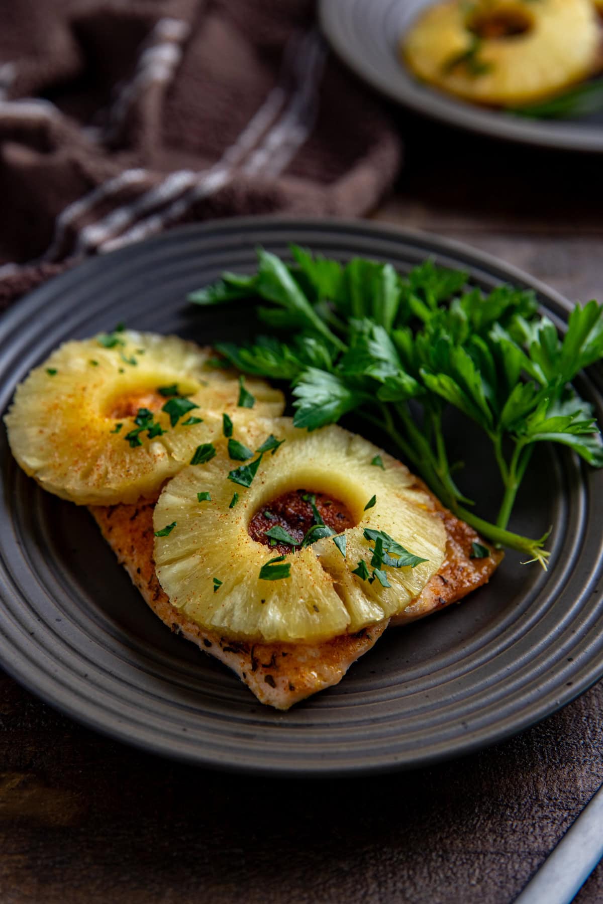 One baked salmon fillet topped with two pineapple rings and sprinkled with fresh cut parsley on a brown plate.