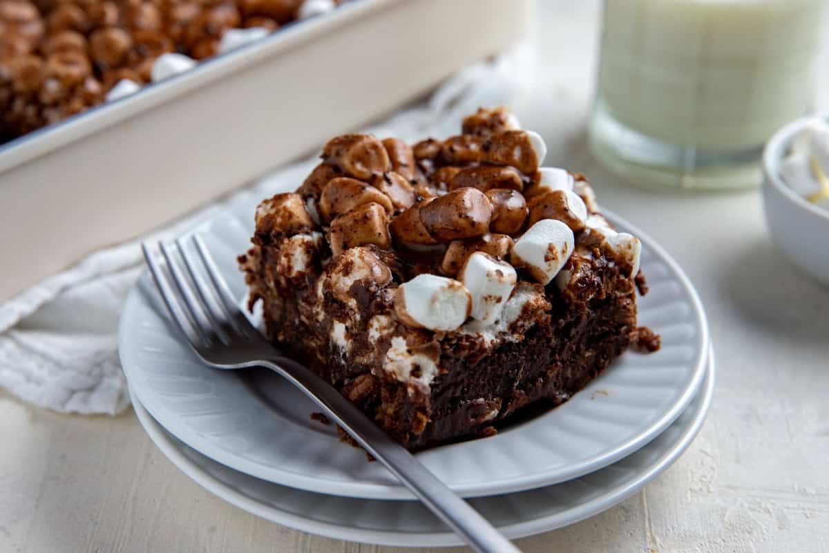One slice of cake on a white plate with a fork next to it. Glass of milk in the background.