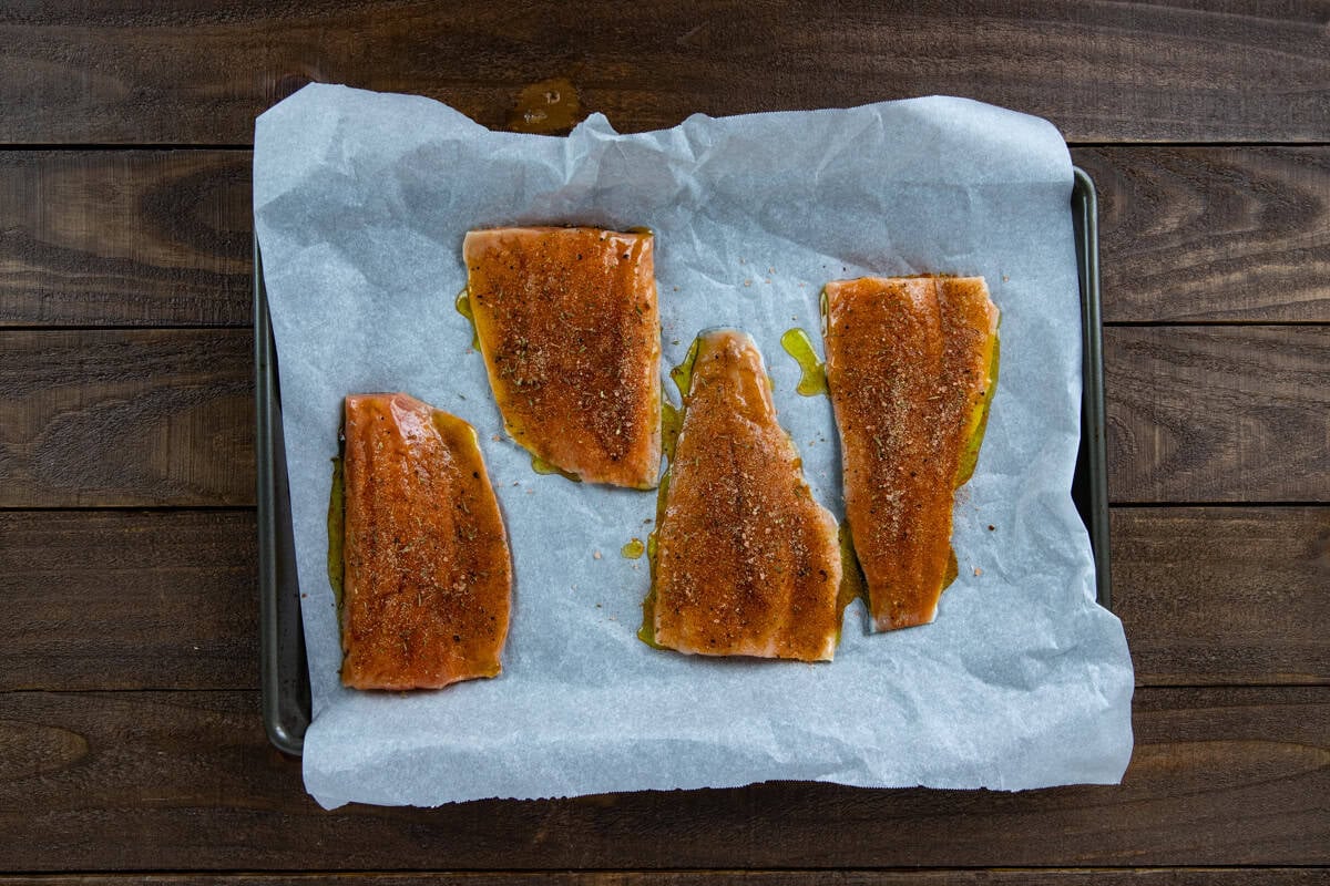 Four uncooked salmon fillets on a parchment lined sheet pan. Topped with honey-olive oil mixture and seasonings.