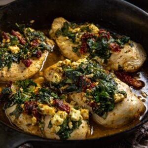 Four baked chicken breasts topped with feta cheese, chopped baby spinach, and sun dried tomatoes in a cast iron skillet.