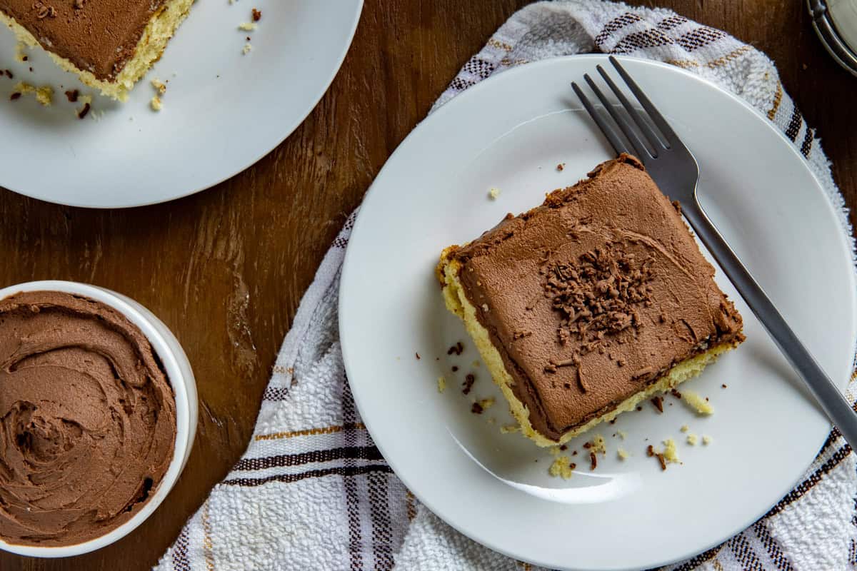 Overhead view of one square slice of frosted cake on a white plate with a fork and a glass of milk in the background.