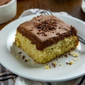A square slice of chocolate frosted vanilla cake on a white plate with a fork.