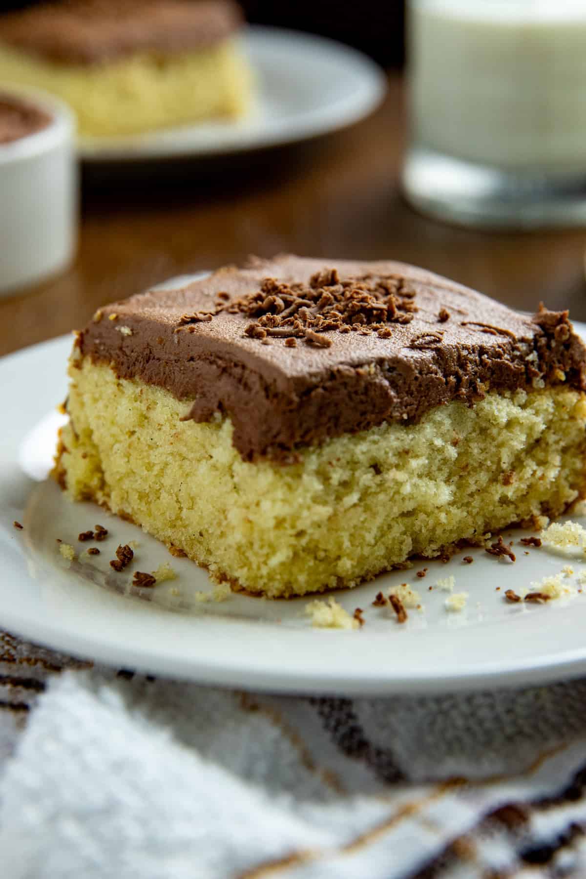 One square slice of frosted cake on a white plate with glass of milk in the background.