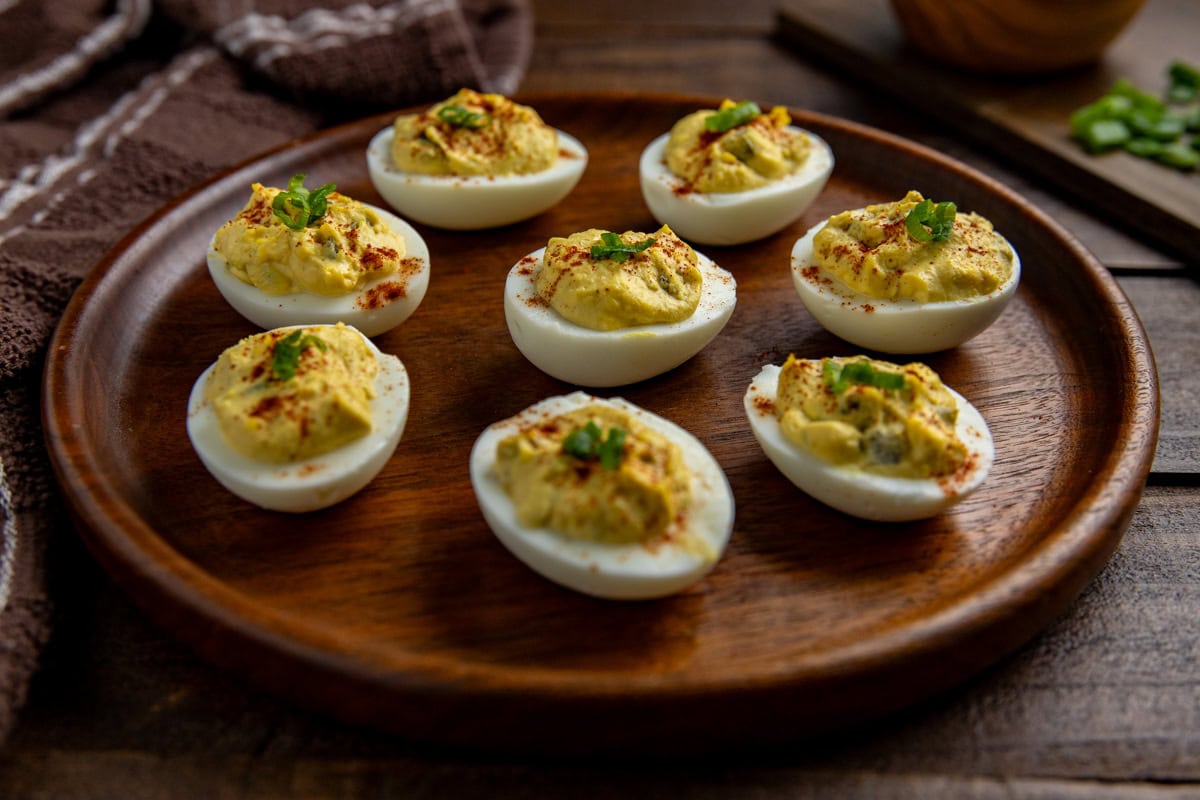 Eight deviled eggs garnished with smoked paprika and sliced green onions on a wooden serving platter.