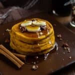 A stack of pumpkin spice pancakes topped with sliced bananas, chopped pecans, and syrup on a plate. Some cinnamon sticks on the plate, too.