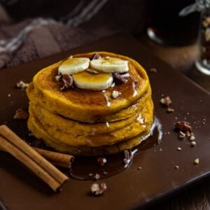 A stack of pumpkin spice pancakes topped with sliced bananas, chopped pecans, and syrup on a plate. Some cinnamon sticks on the plate, too.