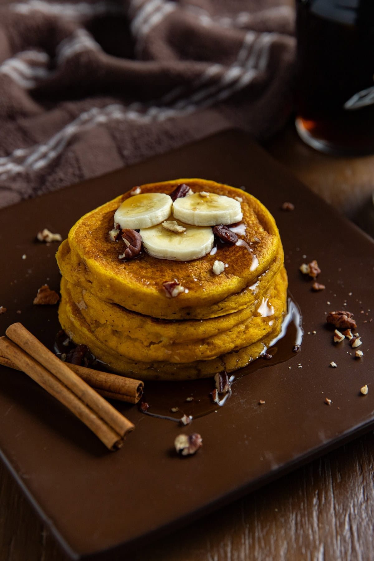 A stack of pumpkin spice pancakes topped with sliced bananas, chopped pecans, and syrup on a plate. Some cinnamon sticks on the plate, too.