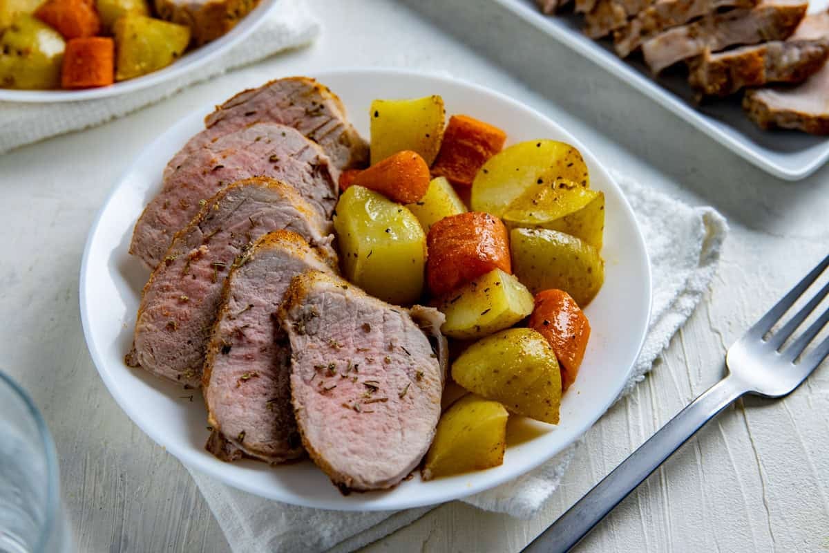 Slices of oven roasted pork tenderloin on a plate with roasted potatoes and carrots.