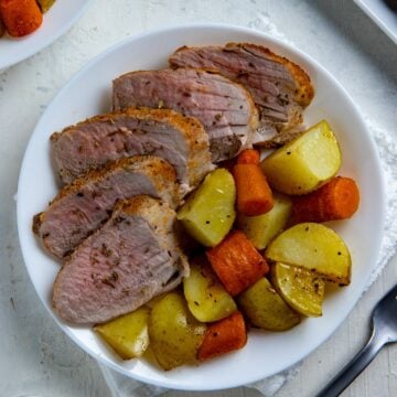 Slices of oven roasted pork tenderloin on a plate with roasted potatoes and carrots.
