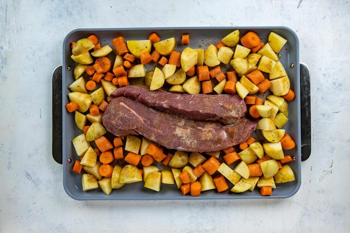 Sheet pan with two uncooked pork tenderloins and chopped potatoes and carrots surrounding it.
