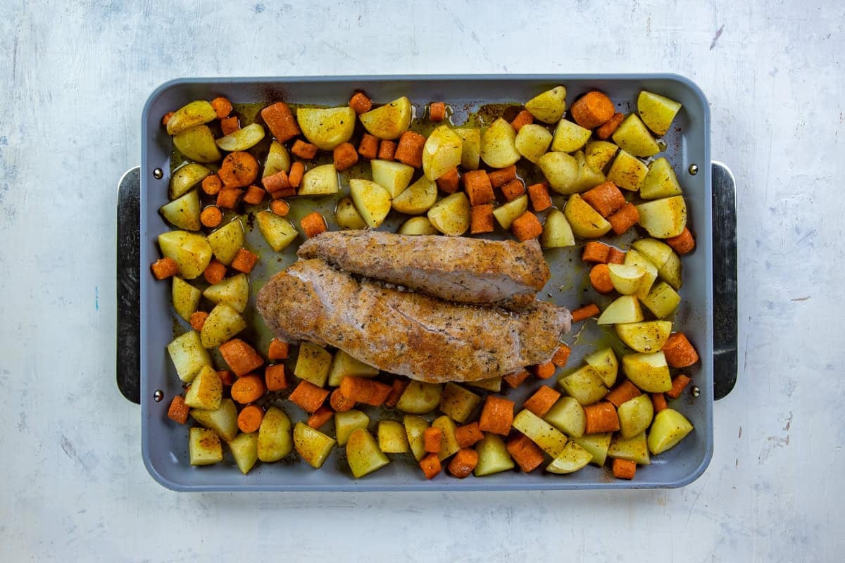 Sheet pan with two roasted pork tenderloins and chopped potatoes and carrots surrounding it.