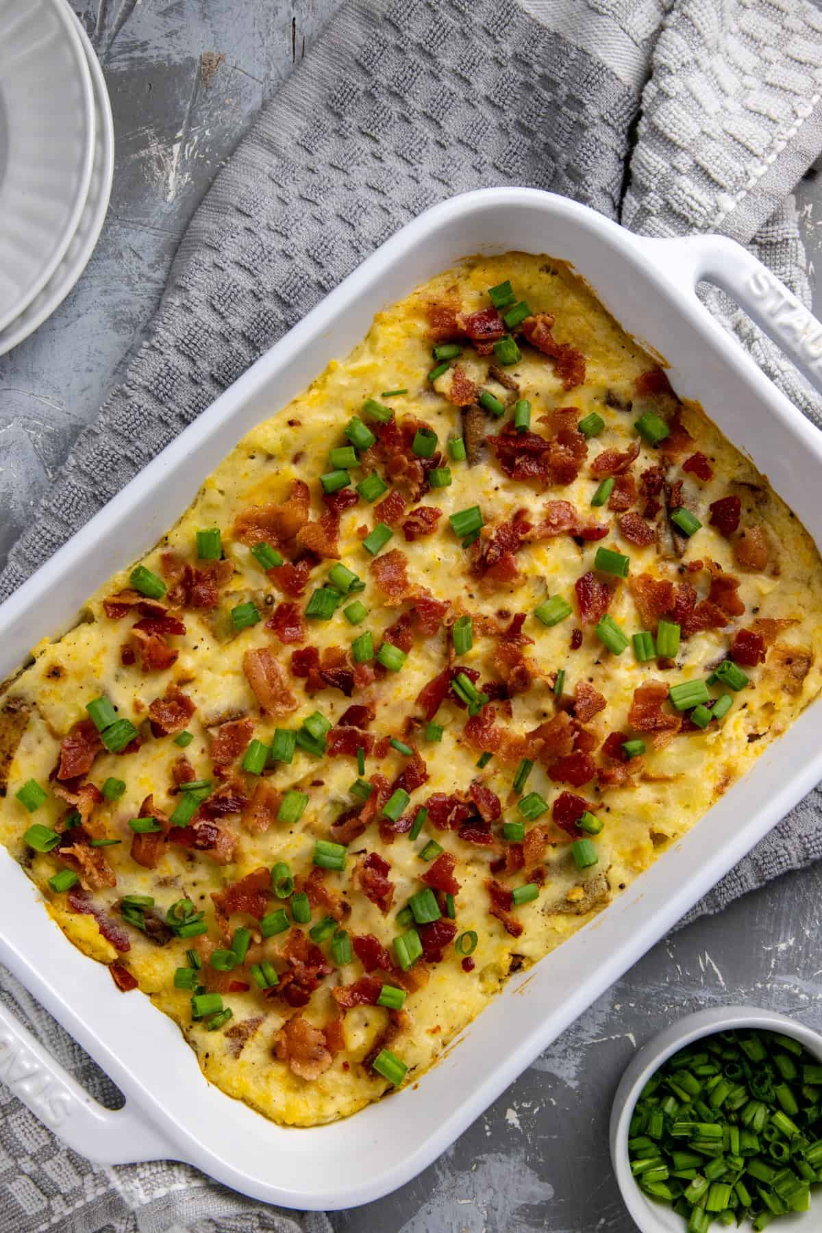 White casserole dish filled with baked mashed potato casserole. Garnished with cooked crumbled bacon and sliced green onions.