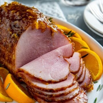 Baked and sliced Ham on a white serving platter with orange slices and rosemary sprigs as garnish.