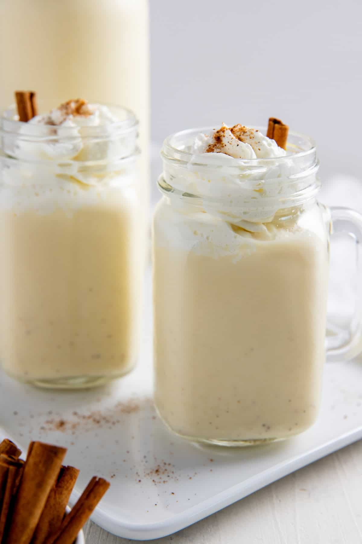 Eggnog in two mason jars topped with whipped cream, nutmeg, and cinnamon sticks.