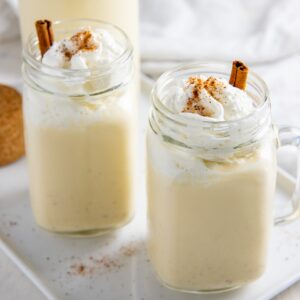 Eggnog in two mason jars topped with whipped cream, nutmeg, and cinnamon sticks.