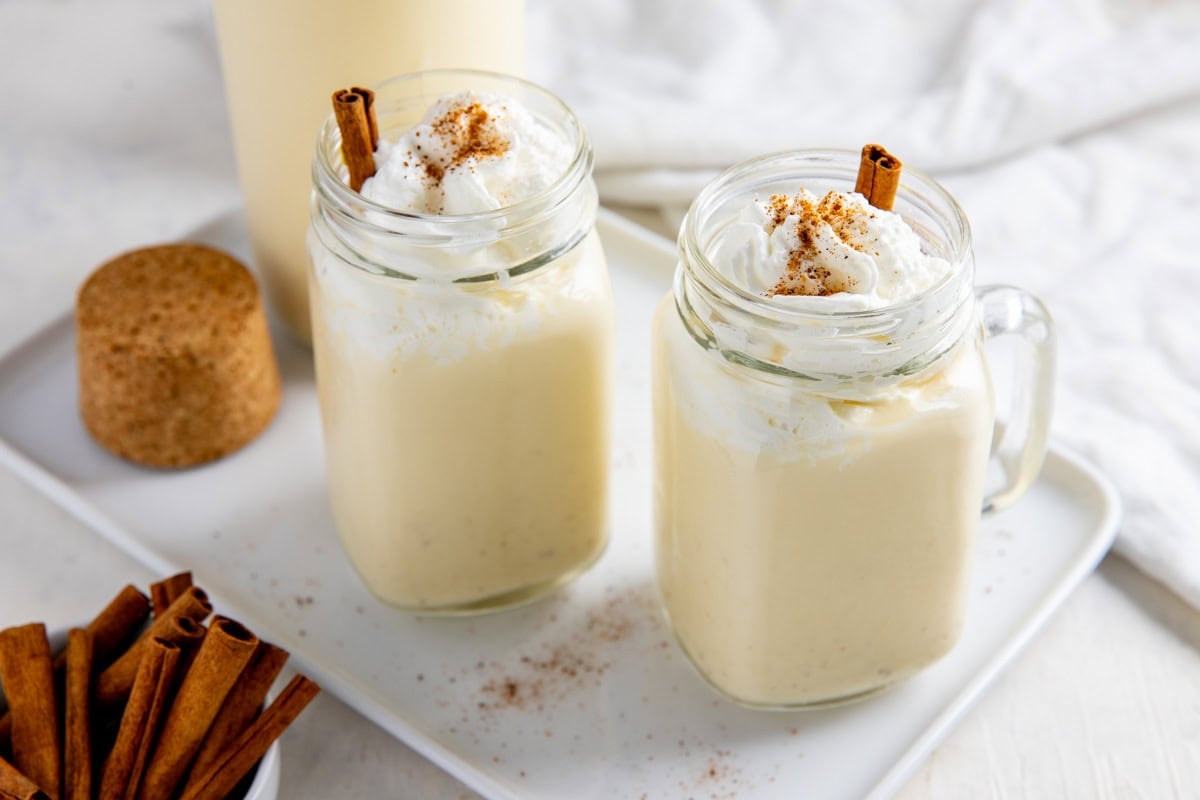 Eggnog in two mason jars topped with whipped cream, nutmeg, and cinnamon sticks.