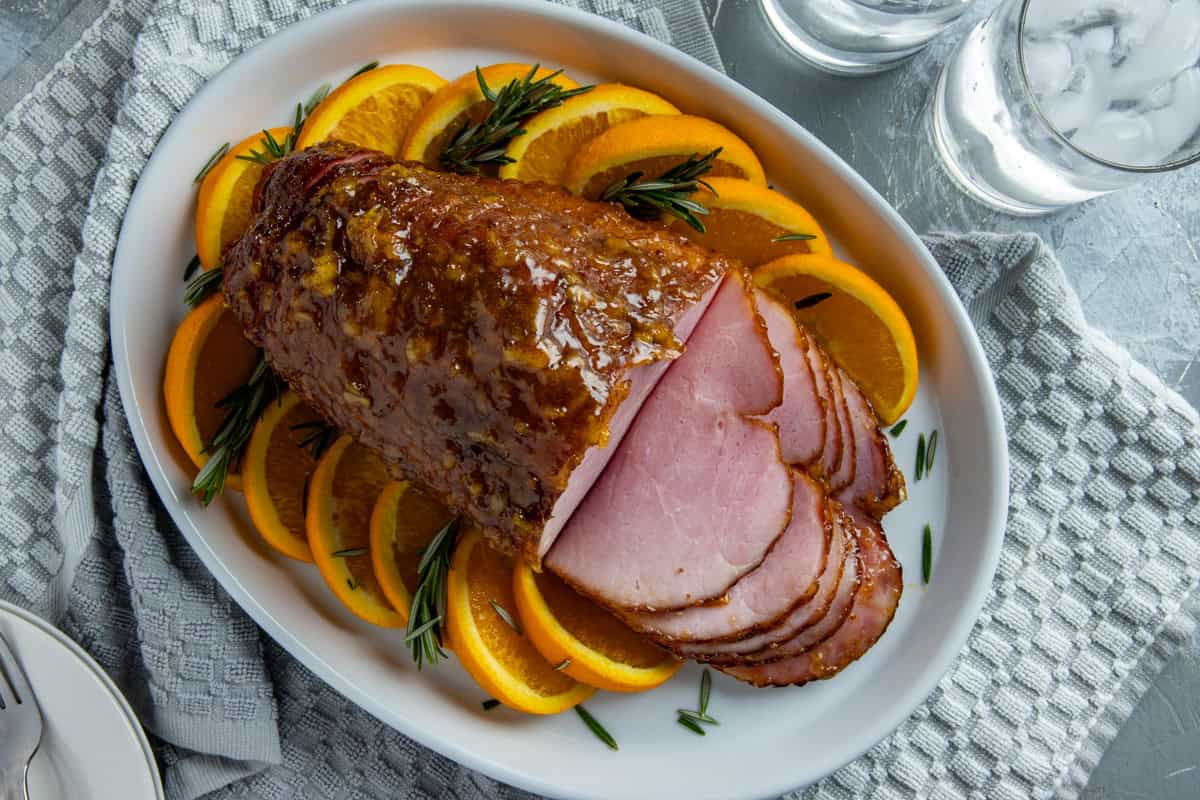 Baked and sliced ham on a white serving platter with orange slices and rosemary sprigs as garnish.