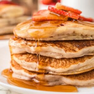 Four pancakes stacked on top of each other with sprup dripped down the sides and sliced strawberries on top.