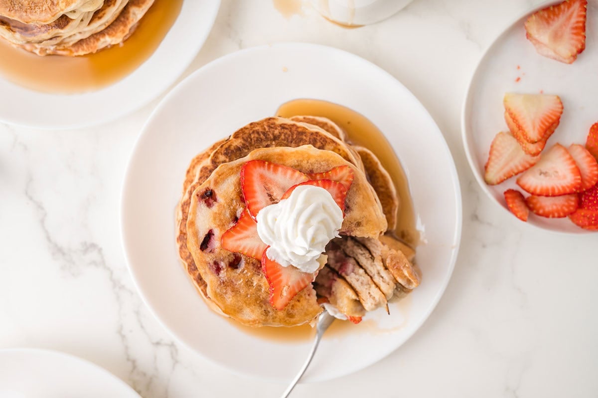 Overhead view of four stacked pancakes with maple syrup flowing down the sides, sliced strawberries and whipped cream on top.