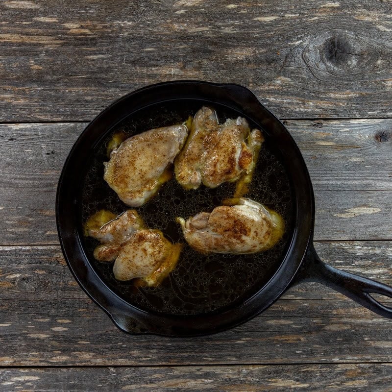Four browned chicken thighs with beef broth and water in a cast iron skillet.