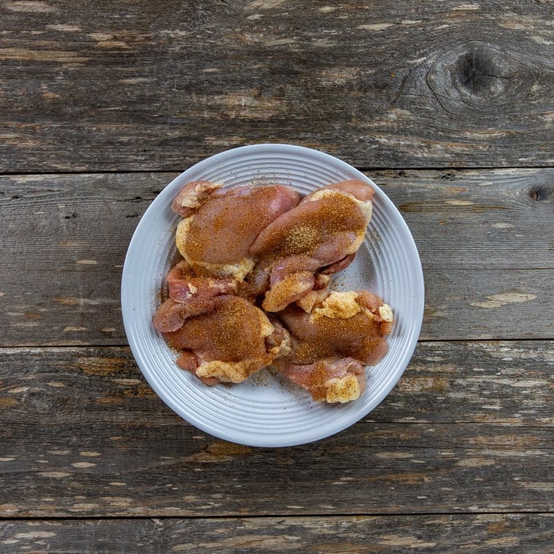 Four raw boneless, skinless chicken thighs seasoned with Creole seasoning on a white plate.