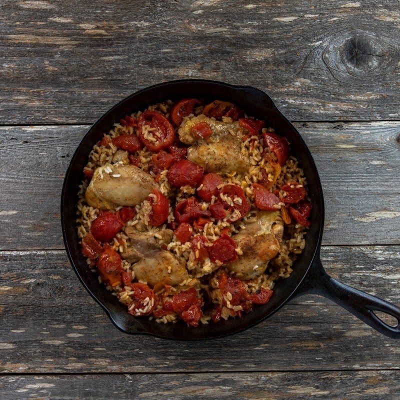 Four browned chicken thighs with beef broth, water, rice, and tomatoes in a cast iron skillet.