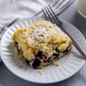 A square slice of blueberry lemon coffee cake with a sprinkling of powdered sugar. On a white plate with a fork.
