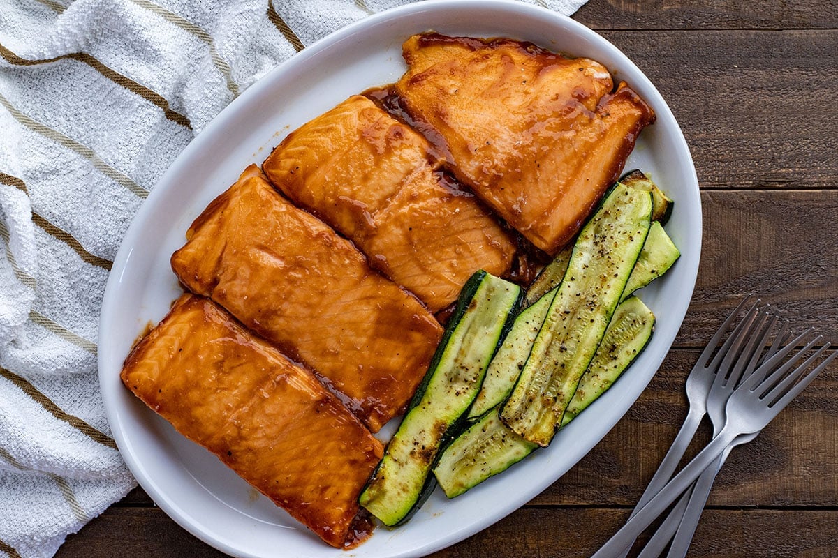 Overhead view of four grilled salmon fillets topped with BBQ sauce on a white platter. Grilled zucchini slices on the side.