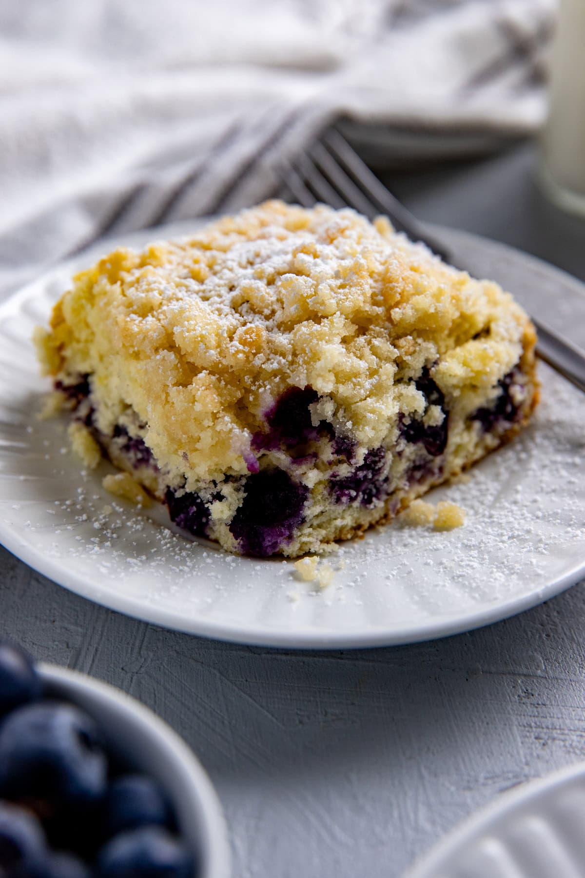 A square slice of blueberry lemon coffee cake with a sprinkling of powdered sugar.  On a white plate with a fork.
